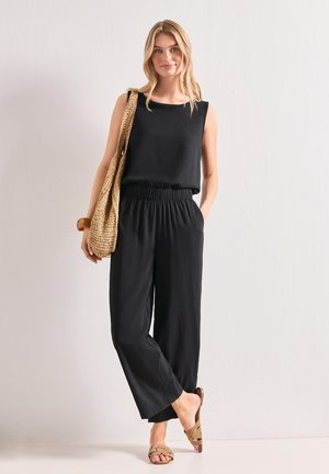 Mujer vestida con top negro sin mangas y pantalones negros sueltos, llevando un bolso de hombro tejido y sandalias color cuero, de pie junto a una pared lisa.