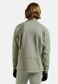 Hellgrüne, langärmelige Jacke mit lockerem Schnitt, hohem Kragen und kontrastierenden Nähten; dazu schwarze Handschuhe.