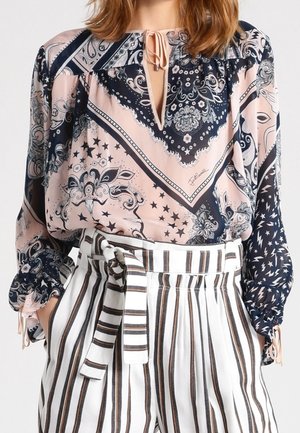 Vrouw die een blouse draagt met een paisley-patroon in roze en marineblauw met ballonmouwen, gecombineerd met een witte broek met hoge taille en verticale strepen in bruin en zwart.