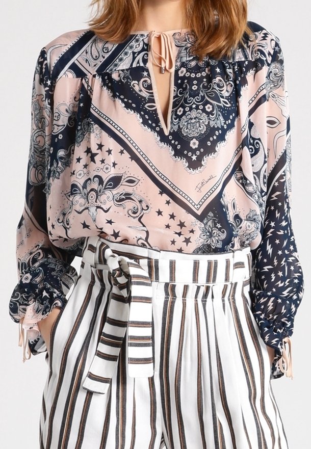 Femme portant un chemisier rose et bleu marine à motif cachemire avec des manches ballon, associé à un pantalon blanc taille haute avec des rayures verticales marron et noires.