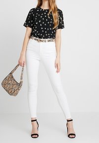 Camisa negra de manga corta con estampado de pájaros blancos, jeans ajustados blancos, bolso de serpiente ysandalias de tacón negras. Atuendo sencillo y moderno.