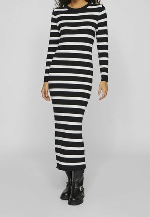 Femme portant une robe en maille à rayures horizontales noires et blanches à manches longues et des bottines noires à boucle, debout devant un fond uni.