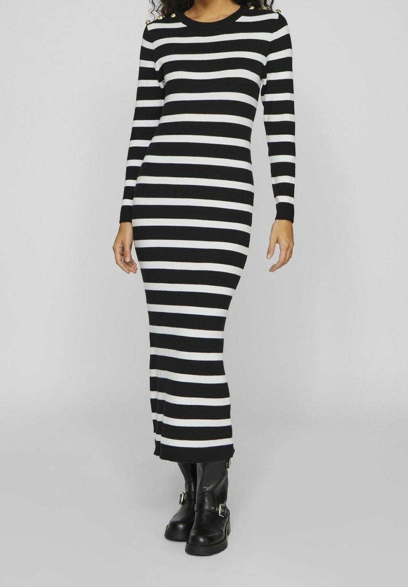 Femme portant une robe en maille à rayures horizontales noires et blanches à manches longues et des bottines noires à boucle, debout devant un fond uni.
