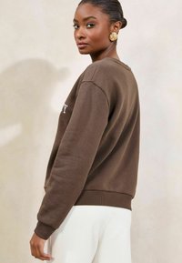 Felpa marrone con collo a giro, polsini e orlo a coste. Presenta testo bianco sul davanti. Il modello indossa pantaloni bianchi e orecchini dorati.