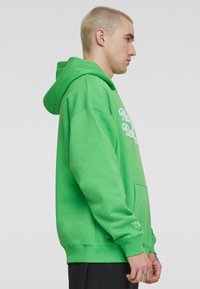 Sudadera de algodón verde con bolsillo canguro, puños acanalados y bordado "New York 93" en la manga. Vista lateral mostrada.