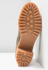 Semelle d'une botte de couleur kaki avec des crampons profonds et des motifs en forme de fleurs, photographiée sur un fond blanc.