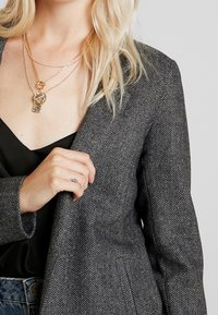 Grau karierter Blazer mit strukturierter Passform, ausgestattet mit einem Schößchen-Revers und strukturierter Stoffoberfläche. Eine mehrschichtige goldene Halskette ist sichtbar.