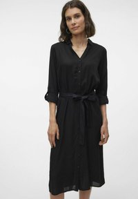 Abito camicia nero con colletto, maniche lunghe arrotolate, chiusura con bottoni e cintura abbinata. Tessuto liscio, taglio al ginocchio, decorazioni minime.