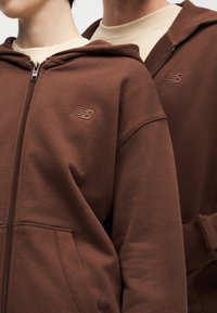 Δύο άτομα φορούν καφέ φούτερ με φερμουάρ και μπεζ πουκάμισα από κάτω, με ένα διακριτικό κεντητό λογότυπο στο στήθος.