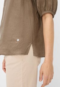 Blusa di lino marrone con maniche a sbuffo e una fessura laterale, impreziosita da un elemento circolare argentato all'orlo. Sono visibili pantaloni beige chiaro.