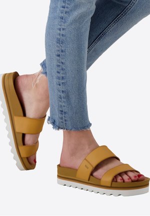 Yellow slide sandals with two wide straps, thick white sole, and textured base. Worn with frayed hem blue jeans, showcasing the footwear.