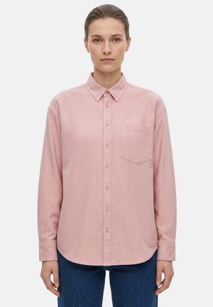 Donna con i capelli legati che indossa una camicia rosa chiaro con bottoni e taschino sul petto e jeans blu, in piedi davanti a uno sfondo semplice.