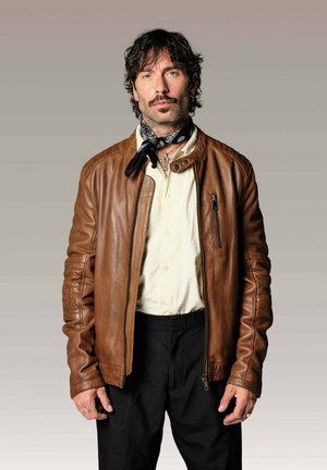 Homme aux cheveux bouclés et moustache portant une veste en cuir marron, une chemise blanche, un pantalon noir et un foulard noir à motifs sur fond gris.