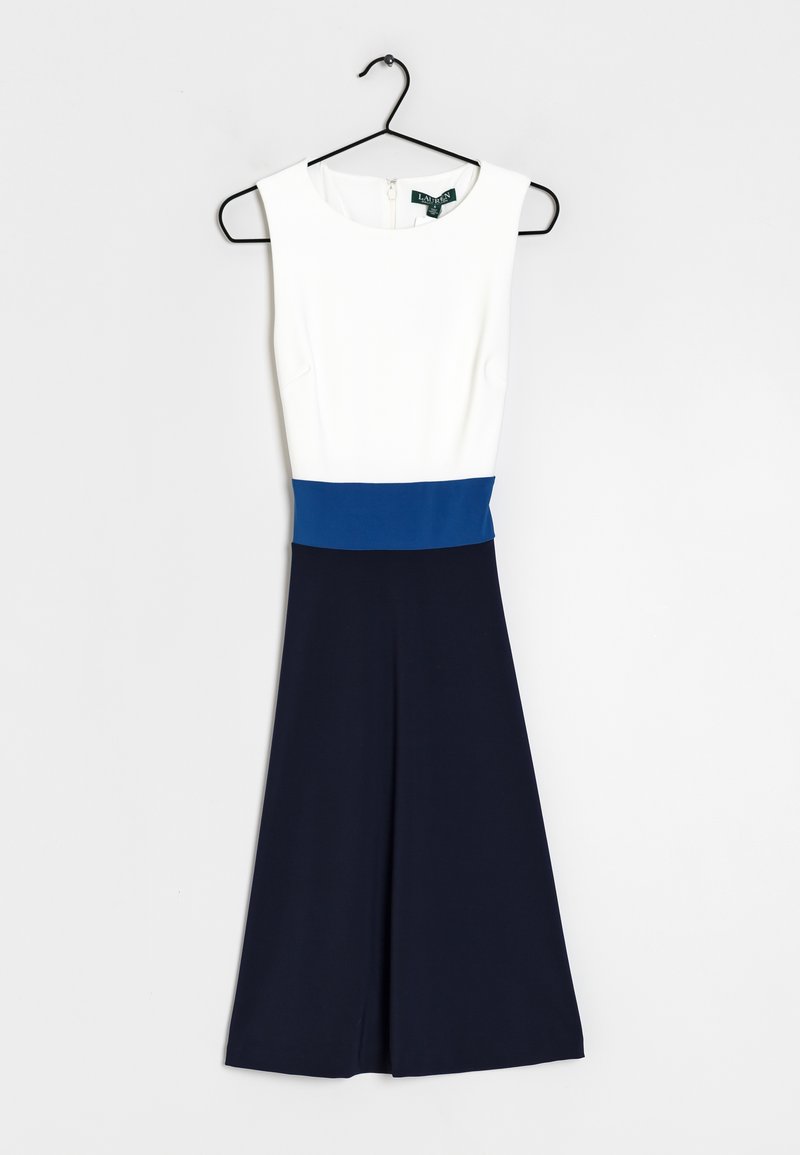 Robe sans manches avec un corsage blanc, une large ceinture bleue et une jupe évasée bleu marine accrochée à un cintre noir contre un mur blanc.