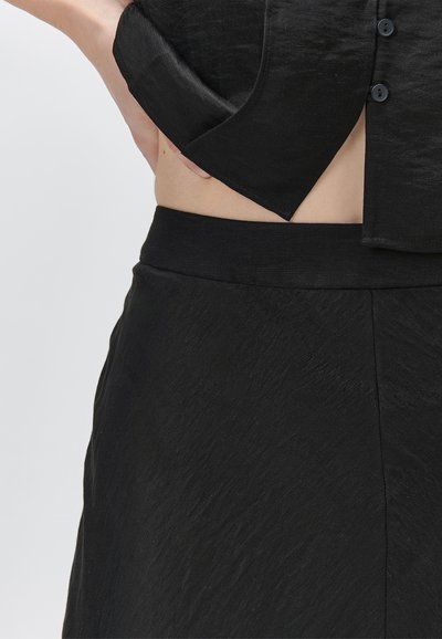 Personne portant un pantalon noir texturé et un haut noir boutonné avec la main glissée dans la poche latérale, montrant une découpe au niveau du ventre.