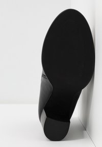Botte cheville en cuir noir montrée par la semelle, inclinée contre un fond blanc en angle.