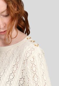 Pull à mailles crème clair avec un motif en dentelle, doté d'une encolure large et de quatre boutons décoratifs en ton or sur l'épaule gauche.