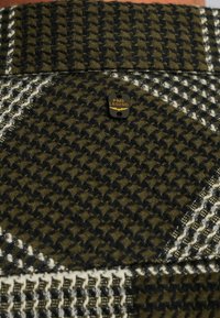 Tissu en laine avec un motif pied-de-poule en vert foncé et crème, présentant une petite étiquette noire avec "PME LEGEND" en lettres dorées.