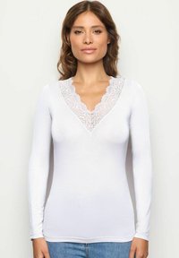 Top bianco a maniche lunghe con scollatura a V bordata in pizzo. Tessuto liscio e design aderente, abbinato a un pantalone in denim casual.
