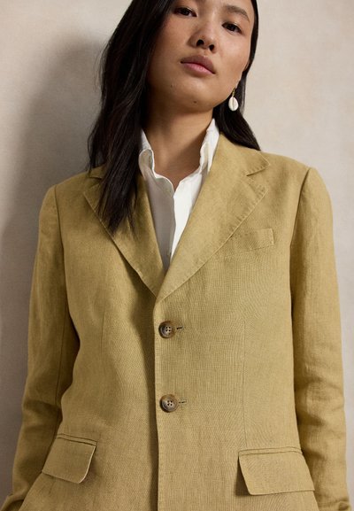 Noor naine, kes kannab sinepikollast jopet valge nööbitava särgi kohal, pikkade tumedate juustega ja ühe rippuva kõrvarõngaga.