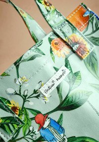 Geantă tip tote cu model floral, pe fundal verde mentă. Prezintă ilustrații cu flori și un personaj purtând o pălărie roșie. Material din bumbac.