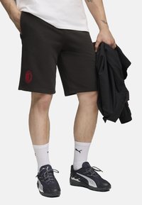 Sorte bomuldsshorts med et rødt broderet logo, knælange, med sidelommer, båret med hvide sokker og mørke sportssko.