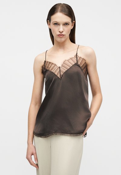 Γυναίκα με ίσια καστανά μαλλιά, που φοράει μια καφέ σατέν καλοκαιρινή μπλούζα με διαφανή δαντέλα στο τελείωμα και ανοιχτό μπεζ παντελόνι, σε απλό φόντο.