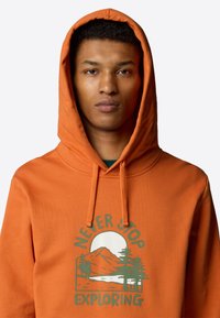 Felpa arancione con un design grafico che presenta montagne, alberi e la scritta "NON SMETTERE MAI DI ESPLORARE" in verde. Tessuto morbido, cappuccio con cordino.
