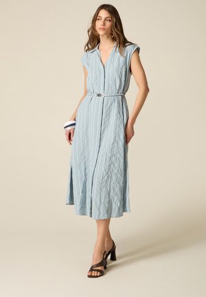 Frau in ärmellosem, hellblau gestreiftem Midikleid mit Gürtel, trägt schwarze Sandalen mit Absatz und weiße Armbänder, steht vor beigem Hintergrund.