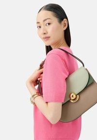 Torebka na ramię z beżową bazą, zielonymi i kremowymi akcentami oraz okrągłym złotym okuciem. Modelka nosi różową bluzkę i złote bransoletki.