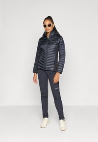 Polstret sort jakke med lynlås og hætte; sorte bukser; hvide sneakers; modellen bærer solbriller; tekstureret finish og kontrasterende paneler.