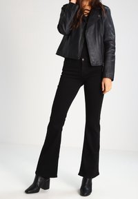 Černá kožená bunda s zipovými kapsami, která je vrstvená přes černý top; spárována s rozšířenými černými džíny a černými botami na podpatku do kotníků.