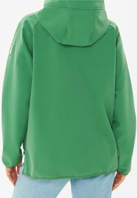 Personne portant une veste à capuche verte vue de dos, associée à un jean bleu clair, les mains détendues le long du corps.