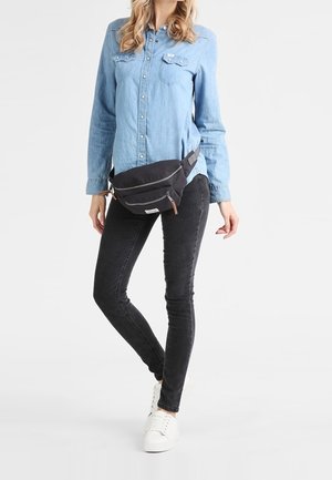 Vrouw draagt een lichtblauw denimoverhemd met knopen, zwarte skinny jeans, witte sneakers en een zwarte heuptas met ritssluitingen.