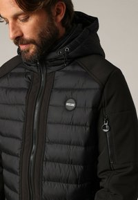 Homme avec une barbe portant une veste matelassée noire Deeluxe avec capuche, fermeture éclair à l'avant, et poche zippée sur la manche gauche, regardant vers le bas.