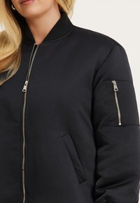 Vrouw in een zwarte bomberjack met zilveren ritsdetails aan de voorkant en op de mouwzak, met lang blond haar zichtbaar.