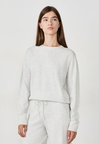 Young woman with long brown hair wearing light gray knit sweater and matching pants, standing against plain background.