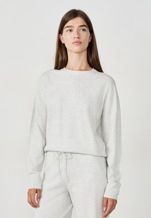 Junge Frau mit langen braunen Haaren, die einen hellgrauen Strickpullover und passende Hose trägt, steht vor einem schlichten Hintergrund.