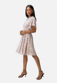 Blumenkleid mit kurzen Ärmeln, pinkem Grundton und grünen, gelben sowie roten Mustern; hat eine taillierte Taille, einen fließenden Rock und schwarze Absatzsandalen.