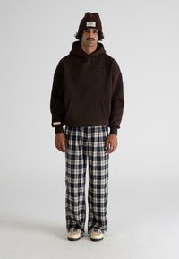 Hombre con un gorro marrón, sudadera marrón oscuro, pantalones a cuadros en blanco y negro, y zapatos beige, de pie contra un fondo claro y liso.