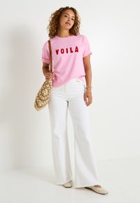 Camiseta de algodón rosa con el logo "VOILÀ" en rojo, combinada con pantalones acampanados blancos. Un bolso de mimbre beige y zapatos claros completan el look.
