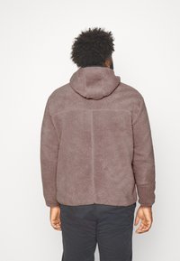 Brun fleecejakke med hætte, der har en tekstureret overflade og en afslappet pasform. Bagsiden viser en central søm og en lige kant.