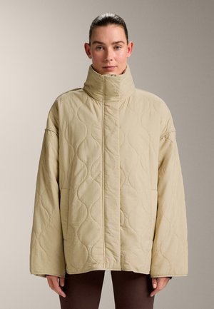 Kvinde med opsat hår iført stor beige quiltet jakke med høj krave, stående foran en ensfarvet baggrund.