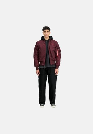 Burgunderfarbene Bomberjacke mit strukturiertem Finish, Kapuzen-Design, Frontreißverschluss und Seitentasche. Kombiniert mit schwarzen Cargo-Hosen und Sneakers.