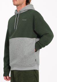Hætte sweatshirt i mørkegrøn og grå, med en kængurulomme, snore og kontrasterende stofsektioner for tekstur og varme.