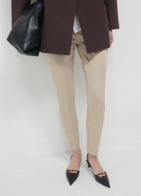 Pantalon beige sur mesure associé à un blazer oversize marron foncé. Sac à main en cuir noir et escarpins à bouts pointus avec un accent doré complètent le look.