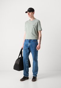 Lys grønn bomulls t-skjorte med liten logo, blå denimjeans, svart caps med hvit søm, svart duffelbag og brune sko.