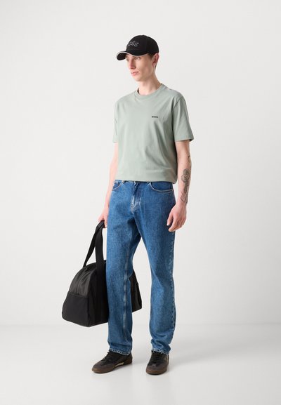 T-shirt en coton vert clair avec un petit logo, jeans en denim bleu, casquette noire avec des coutures blanches, sac de sport noir et chaussures marron.