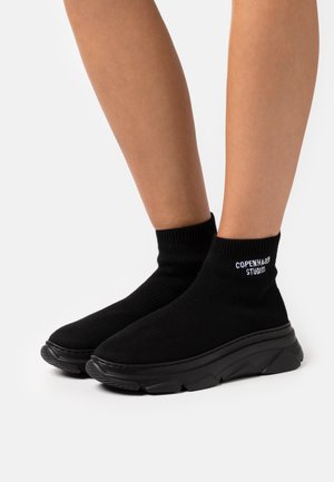 Baskets chaussettes noires à semelles épaisses avec le texte « Copenhagen Studios » portées sur des jambes nues sur un fond blanc uni.