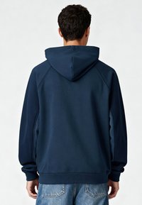 Sweat à capuche bleu marine avec capuche à cordon, manches raglan et poignets côtelés. Présente des panneaux contrastants sur les côtés et une texture lisse.